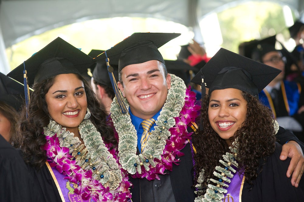 Three political science majors wearing leis and caps and gowns smile.